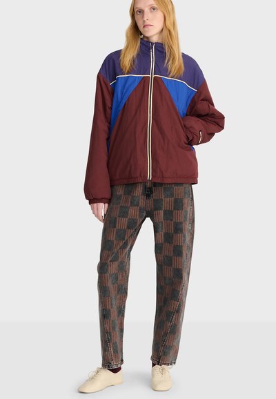 Femme portant une veste zippée bordeaux, bleue et violette, un pantalon à carreaux marron et noir, et des chaussures beiges, debout devant un fond uni.