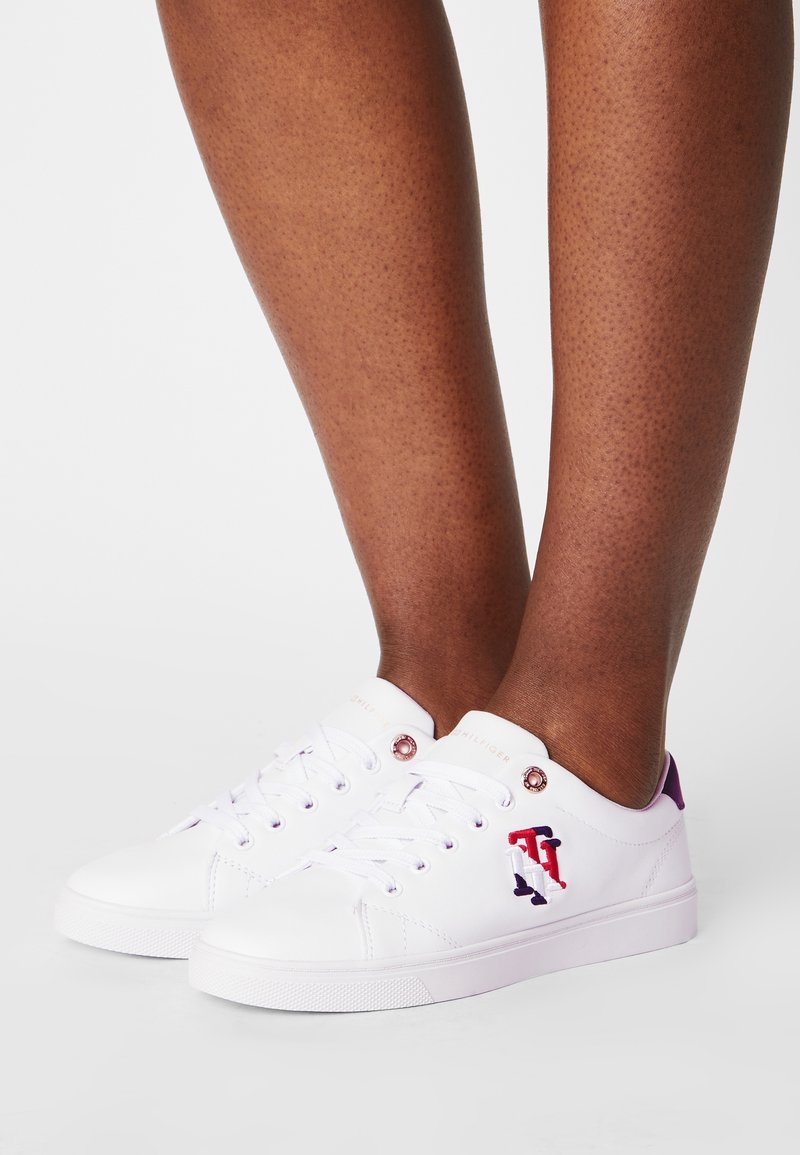 White canvas sneakers with purple accents, round toe, flat rubber sole, and embroidered logo on the side. Eyelets and laces included.