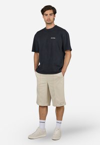 Zwarte katoenen T-shirt met korte mouwen en een wit logo, gecombineerd met beige shorts met dubbele stiksels en een merklabel. Witte sneakers.