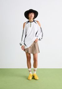 Witte langlopende blouse met uitsneden op de schouders, zwarte strepen en een kleurrijke trekkoord; gecombineerd met een beige plissé rok en gele schoenen.