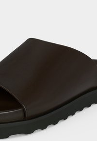 Sandale à enfiler marron foncé avec tige large en cuir et semelle en caoutchouc noire rainurée, montrée en gros plan sur un fond clair.