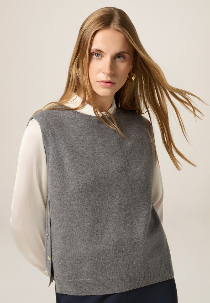 Jersey sin mangas gris de textura suave, con cuello redondo y detalles de botones laterales, combinado con una blusa blanca.