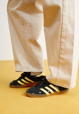 Person trägt beige Hose und schwarze Adidas Gazelle Sneakers mit goldenen Streifen und kleinen rosa Blumenmustern auf einem gelben Boden.