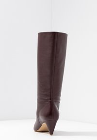 Bottine bordeaux haute jusqu'au genou en cuir lisse, avec un talon effilé et des coutures discrètes. Pas de ferrures ni de motifs visibles.