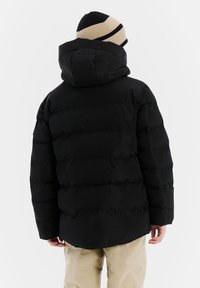 Zwarte pufferjack met capuchon, met doorgestikte delen en een gladde textuur. Gemaakt met een beige en zwart gestreepte gebreide beanie.