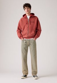 Sudadera roja con cremallera, gran bolsillo frontal y logo, combinada con jeans verde claro y zapatillas en blanco y negro. Tela suave y casual.