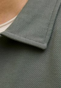 Koszula z kołnierzykiem w przytłumionym odcieniu zieleni, wykonana z fakturowanej tkaniny. Kołnierz jest płaski z starannie wykonanym przeszyciem, ukazując subtelny wzór w jodełkę.