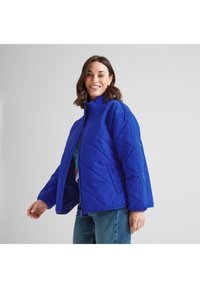 Smiling woman wearing a bright blue quilted jacket and jeans, standing against a plain light gray background.