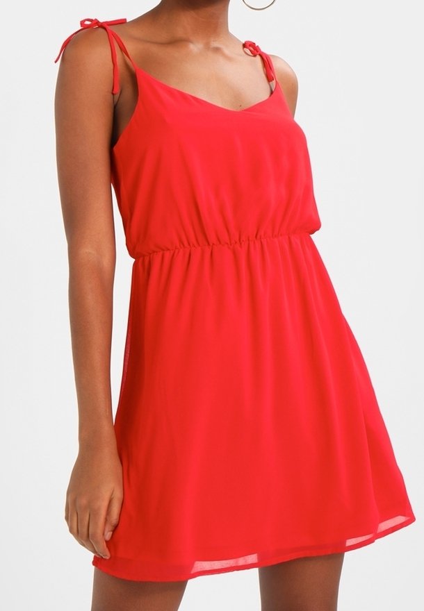Robe rouge sans manches avec bretelles nouées aux épaules, taille froncée et jupe fluide portée par une personne avec des boucles d'oreilles créoles.