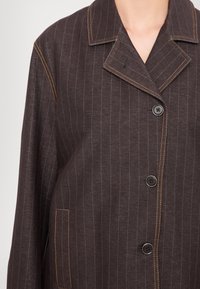 Veste marron foncé à fines rayures avec surpiqûres orange, col cranté, trois boutons noirs et une poche avant côté gauche.