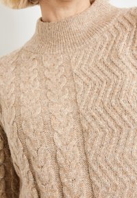 Pull en tricot beige clair avec un col montant, un motif en cable knit d'un côté et un motif chevron de l'autre. Texture douce.