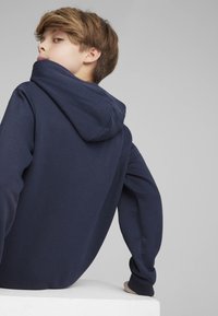 Granatowa bluza z kapturem o miękkiej fakturze, z dużym kapturem i prążkowanymi mankietami. Widok z tyłu ukazuje luźny krój i minimalną ilość szwów.