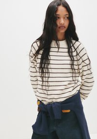 Cremelanger Pullover mit dunklen Streifen, strukturiertem Rippdesign und lockerer Passform. Dunkelblauer Pullover um die Taille über Jeans gebunden.
