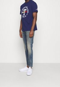 Mężczyzna ubrany w granatową koszulkę Tommy Jeans, wyblakłe niebieskie dżinsy i białe trampki, stojący na tle jednolitego jasnego tła.