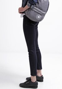 Mochila de lona gris con un logo circular, combinada con jeans ajustados oscuros y zapatillas deportivas negras, enfocándose en un atuendo casual.