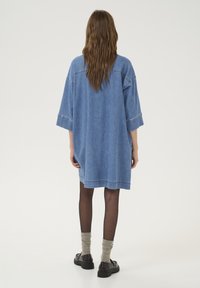 Denim shirt dress, loose fit, mid-thigh length, short sleeves, light blue color, featuring double-stitched seams, worn with dark tights and shoes.