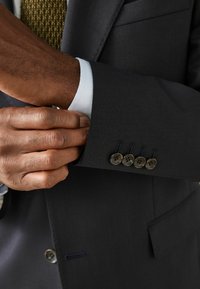 Dunkelgraue Anzugsjacke mit weißem Manschettenende und fünf dunklen Knöpfen am Ärmel. Hand, die die Manschette justiert und den strukturierten Stoff zeigt.