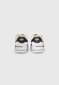 Para białych sneakersów Nike z czarnymi panelami na piętach i beżowymi akcentami, widziana z tyłu na jednolitym białym tle.