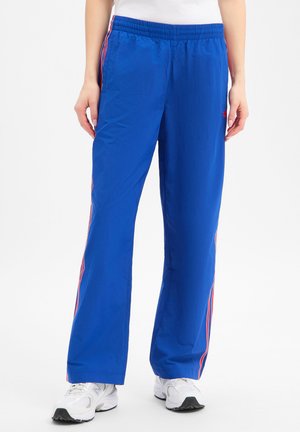 Personne portant un pantalon de survêtement bleu vif avec des bandes latérales rouges et blanches et des baskets blanches, debout devant un fond blanc.
