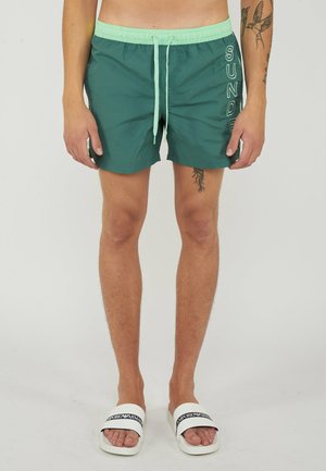 Man draagt groene zwembroek met lichtgroene tailleband en witte koord, witte slippers en zichtbare tattoos op armen en benen.