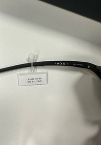 Gafas de sol negras con una montura flexible, que presentan marcas CE grabadas, número de modelo y una etiqueta que indica "NO PARA VENTA" adjunta.