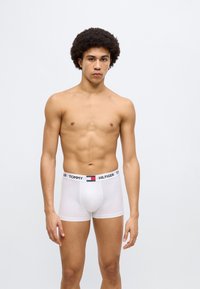Άσπρα βαμβακερά ανδρικά εσώρουχα boxer με μαύρο και πολύχρωμο ελαστικό ζωνάκι "TOMMY HILFIGER", εφαρμοστή σχεδίαση, που φοριούνται από έναν όρθιο άνθρωπο.