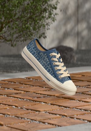 Basket en toile bleue avec lacets blancs et semelle en caoutchouc flottant au-dessus de carreaux en bois en extérieur, avec un mur flou et un feuillage vert en arrière-plan.