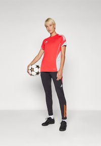 Rød og grå sportsskjorte med korte ermer, parret med sorte bukser med oransje striper. Personen holder en fotball.