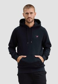 Mężczyzna z krótkimi włosami i brodą, ubrany w czarną bluzę z kapturem z małym różowym logo przedstawiającym skrzyżowane kije do krykieta i piłkę, ręce w przedniej kieszeni.