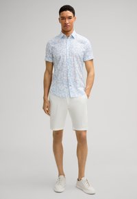 Short-sleeve light blue shirt with floral pattern, paired with white shorts. Model wears white sneakers. Simple design, relaxed fit.