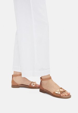 Nohy v béžových sandálech na ploché podrážce s pásky kolem kotníků a kovovými geometrickými ozdobami, kombinované s bílými kalhotami s širokými nohavicemi.