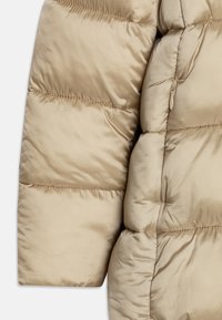 Sezione di giacca trapuntata beige con tessuto liscio e lucido, motivo a rombi, cuciture visibili e una tasca con zip nascosta.