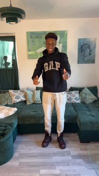 Mladić se smiješi i pokazuje prema naprijed, nosi crnu GAP majicu s kapuljačom, bež hlače i čizme, stoji u dnevnoj sobi s zelenom sofom i zidnom umjetnošću.