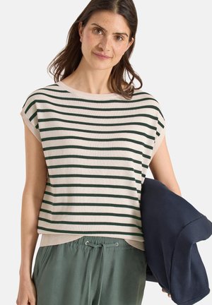 Femme aux cheveux bruns mi-longs portant un haut sans manches beige et vert rayé ainsi qu’un pantalon vert, tenant une veste bleu marine dans la main droite.