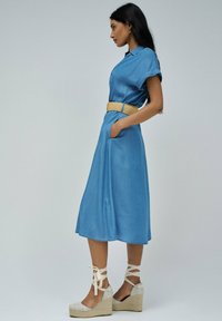 Modra denim obleka s kratkimi rokavi, pasom in žepi. Model nosi bež wedge sandale z vezalkami okoli gležnjev. Gladka tekstura in sproščen dizajn.