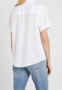 Chemise blanche en lin à manches courtes avec un ourlet arrondi, dotée d'un empiècement au dos et de manches retroussées. Associée à un jean bleu clair.