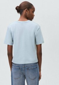 Žena s vlasy do drdolu, oblečená v světle modré košili s krátkým rukávem a modrých džínách, viděná zezadu na neutrálním světle pozadí.