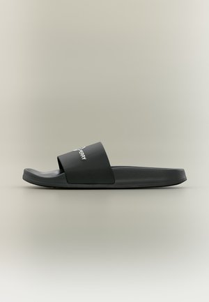 Black slip-on sandal with a wide strap showing partial white brand text, and a contoured footbed on a neutral background.