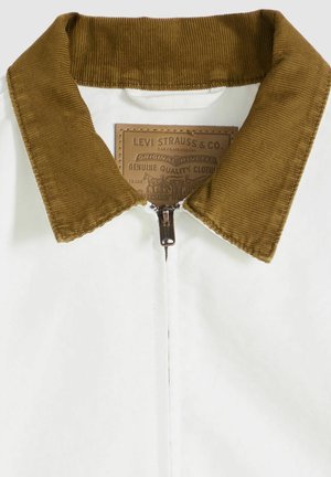 Valkoinen vetoketjullinen takki, jossa on ruskea corduroy-kaulus. Mukana ruskea merkki, jossa brändin nimi ja yksityiskohdat. Sileä pinta, rento muotoilu.