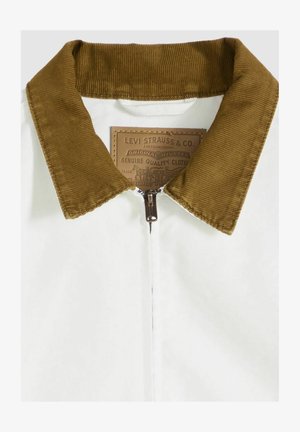 Chaqueta blanca con cremallera completa y cuello de pana marrón. Incluye una etiqueta marrón con el nombre de la marca y detalles. Textura suave, diseño informal.