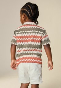 Junges Kind mit geflochtenem Haar, das ein rot-weiß-grün gezacktes Hemd und hellfarbene Shorts trägt, mit dem Rücken zur Kamera gewandt.