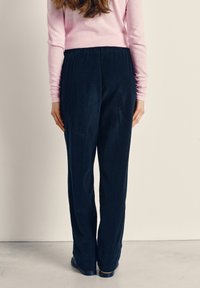 Moteris dėvi rožinę ilgomis rankovėmis palaidinę ir tamsiai mėlynas platėjantį korduroio kelnes, stovi priešais paprastą sieną, atsukusi nugarą.