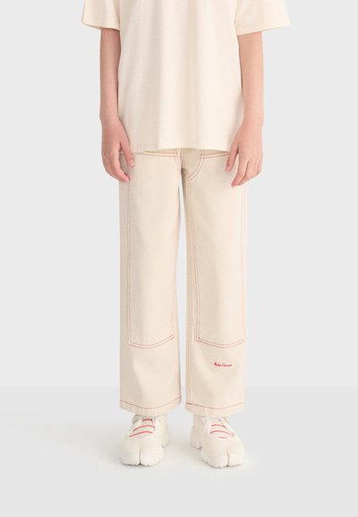 Enfant portant un pantalon large crème avec des surpiqûres rouges et des baskets blanches à bout fendu, debout les mains détendues le long du corps.