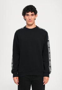 Melna sporta krekla, kas izgatavots no mīkstas auduma, ar garām piedurknēm, uz kurām ir baltas logo akcenti garām plecu līnijām un elastīgām aprocēm. Apaļa kakla izgriezuma forma.