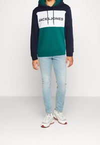 Sudadera bloqueada en verde y marino con el texto "JACK & JONES", combinada con un par de jeans de color azul claro y zapatillas blancas con detalles rojos.