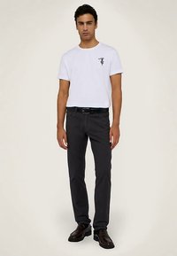 Bílé tričko s grafickou potiskem, tmavě šedé kalhoty, černý pásek a tmavé boty. Model stojí vzpřímeně proti neutrálnímu pozadí.