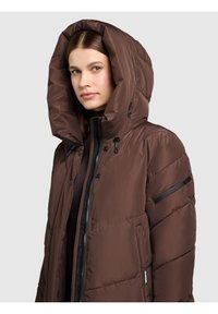 Brązowa puchowa kurtka z dużym kapturem, przednim zamkiem błyskawicznym i detalami na guziki. Posiada kieszeń na rękawie na zamek błyskawiczny, gładką teksturę i pikowany wzór.
