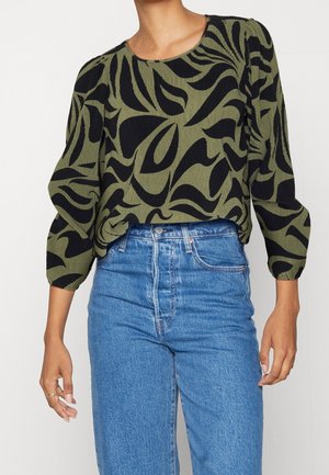 Žena v olivově zelené blůze s černým abstraktním vzorem a nafouklými 3/4 rukávy, doplněná o modré džíny s vysokým pasem.