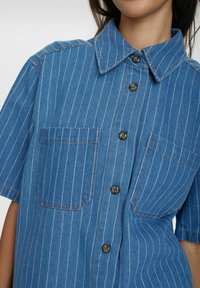 Blå denimskjorta med korta ärmar, vita vertikala ränder, två bröstfickor och bruna knappar. Har en klassisk krage och en strukturerad design.