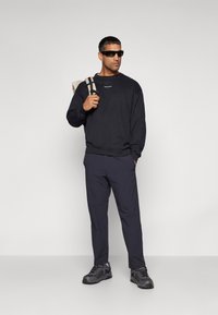 Felpa nera con testo bianco minimalista, pantaloni blu scuro, sneakers scure e occhiali da sole; presenta uno zaino con tracolla a fantasia.